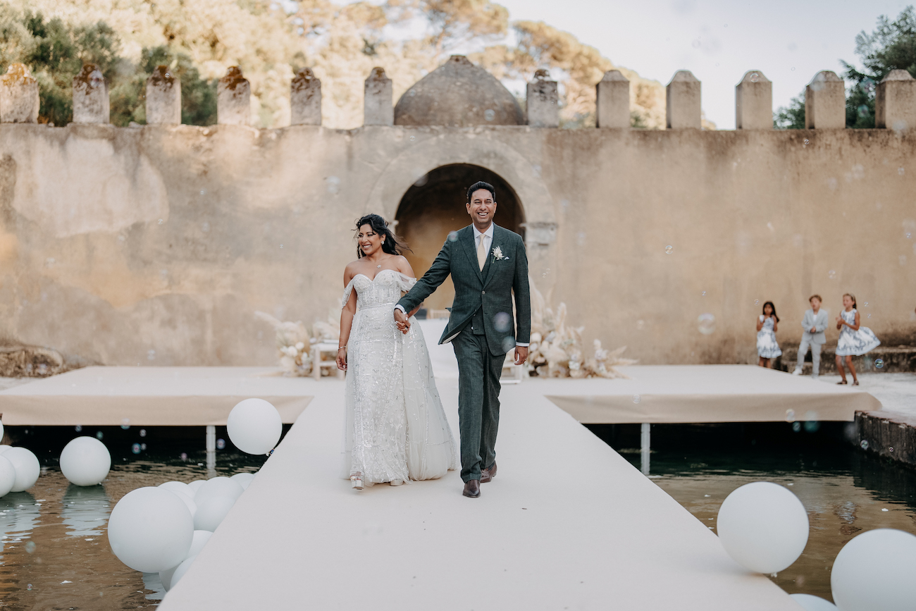 A bride and groom walk hand in hand down an outdoor aisle, smiling, with a stone wall and arches in the background and white balloons along the walkway.