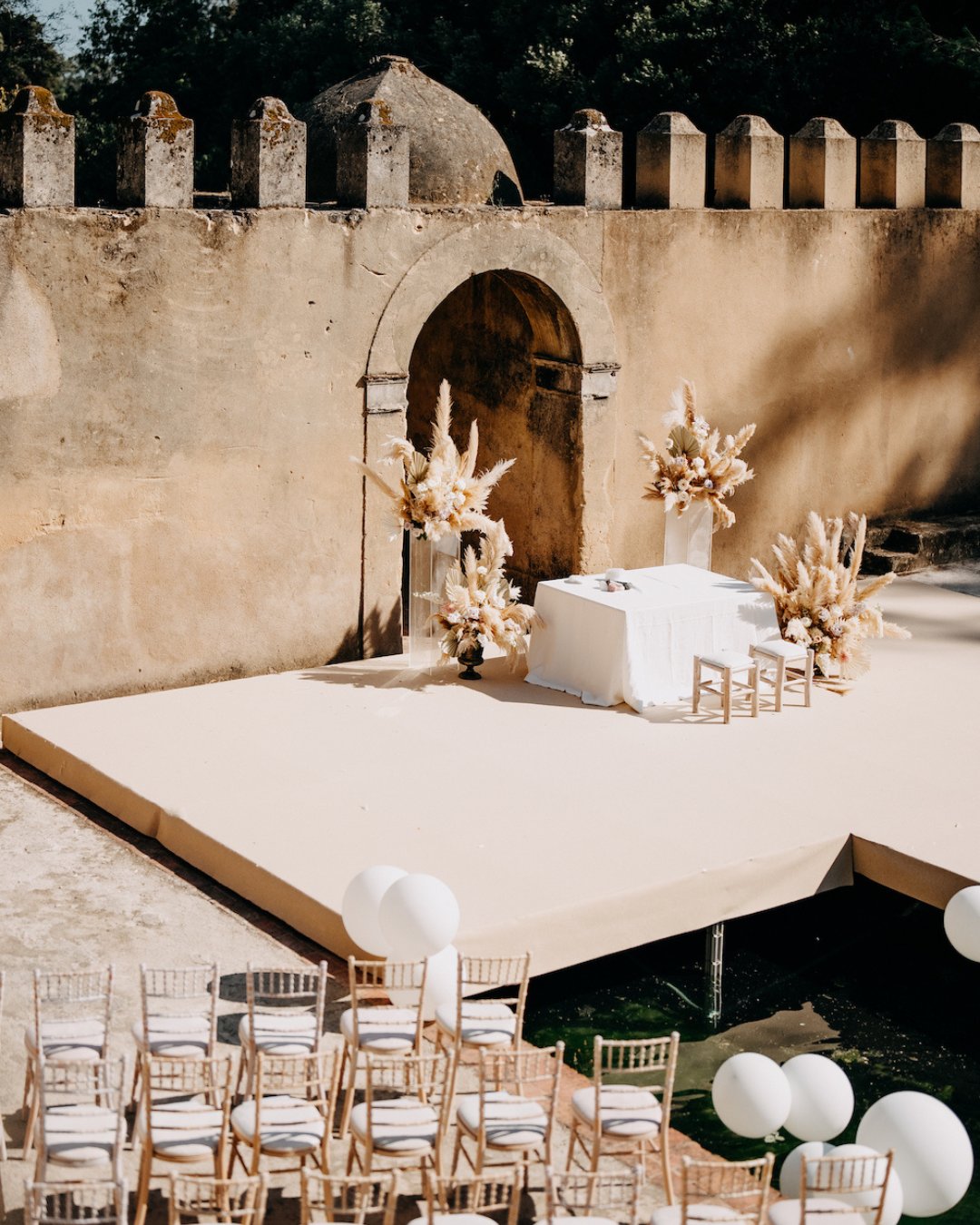 A wedding ceremony setup with a decorated table and chairs on a raised platform in front of an old stone wall, with white balloons and rows of chairs arranged for guests.