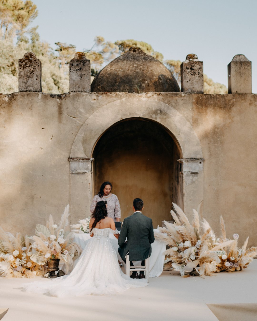 A couple in wedding attire sits facing an officiant in front of an old stone archway, surrounded by neutral floral arrangements.