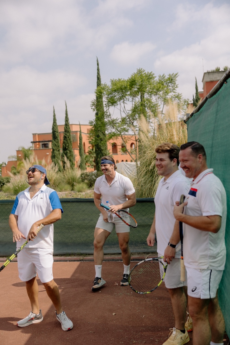 Four men in white tennis outfits stand and laugh on a tennis court, holding rackets, with greenery and a building in the background.