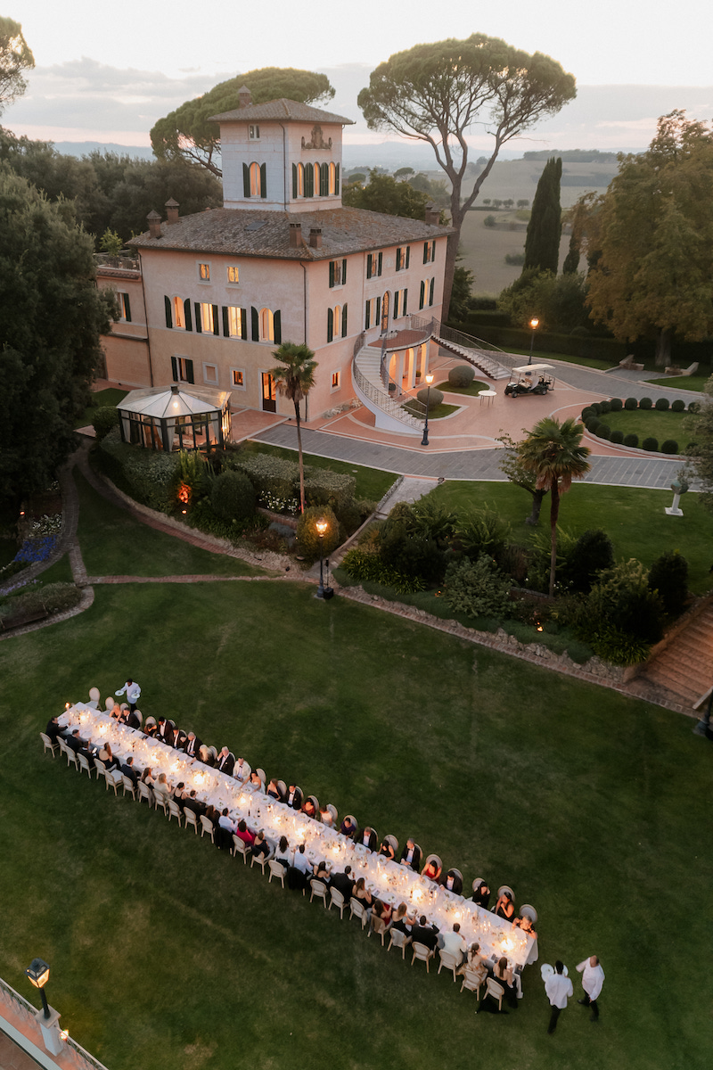 A long dining table with many people seated outdoors on a lawn, in front of a large villa at dusk, with staff serving food.