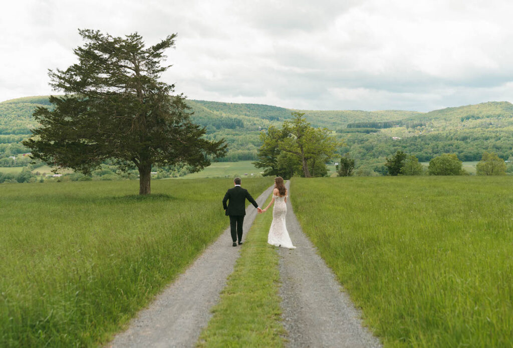 A bride and groom walk hand in hand down a gravel path through a green field, with hills and trees in the background under a cloudy sky.