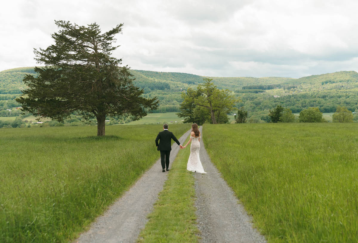 A bride and groom walk hand in hand down a gravel path through a green field, with hills and trees in the background under a cloudy sky.
