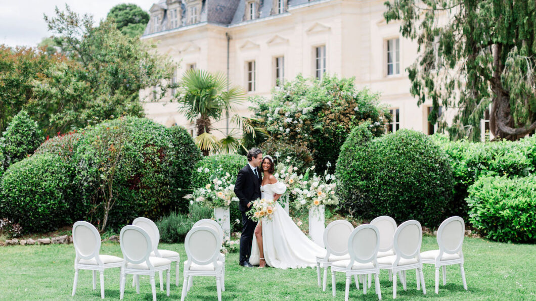 A bride and groom stand together in front of floral arrangements with empty chairs arranged on a lawn, outside a large, elegant building.