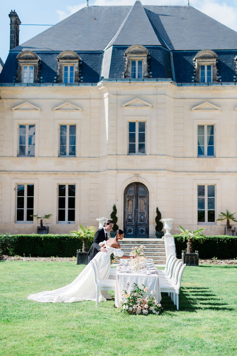 A bride and groom stand beside a decorated outdoor dining table in front of a large, elegant chateau on a sunny day.