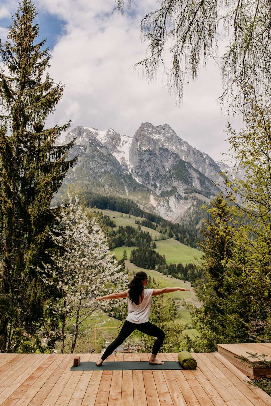 A person practices yoga in warrior pose on a wooden deck, surrounded by trees, with snow-capped mountains in the background.