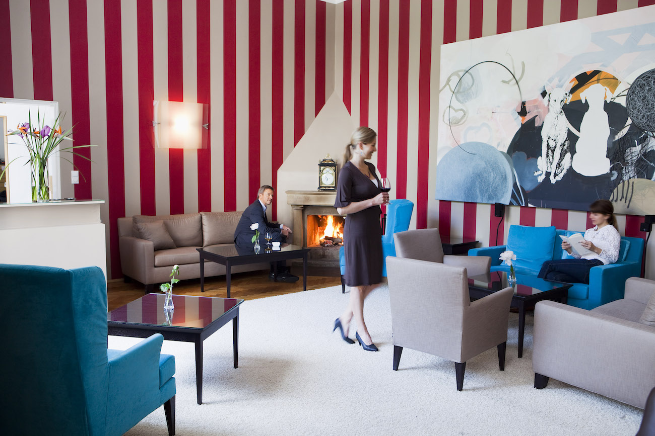 Three people occupy a modern lounge with red and white striped walls, abstract artwork, and a lit fireplace; two are seated and one is standing.