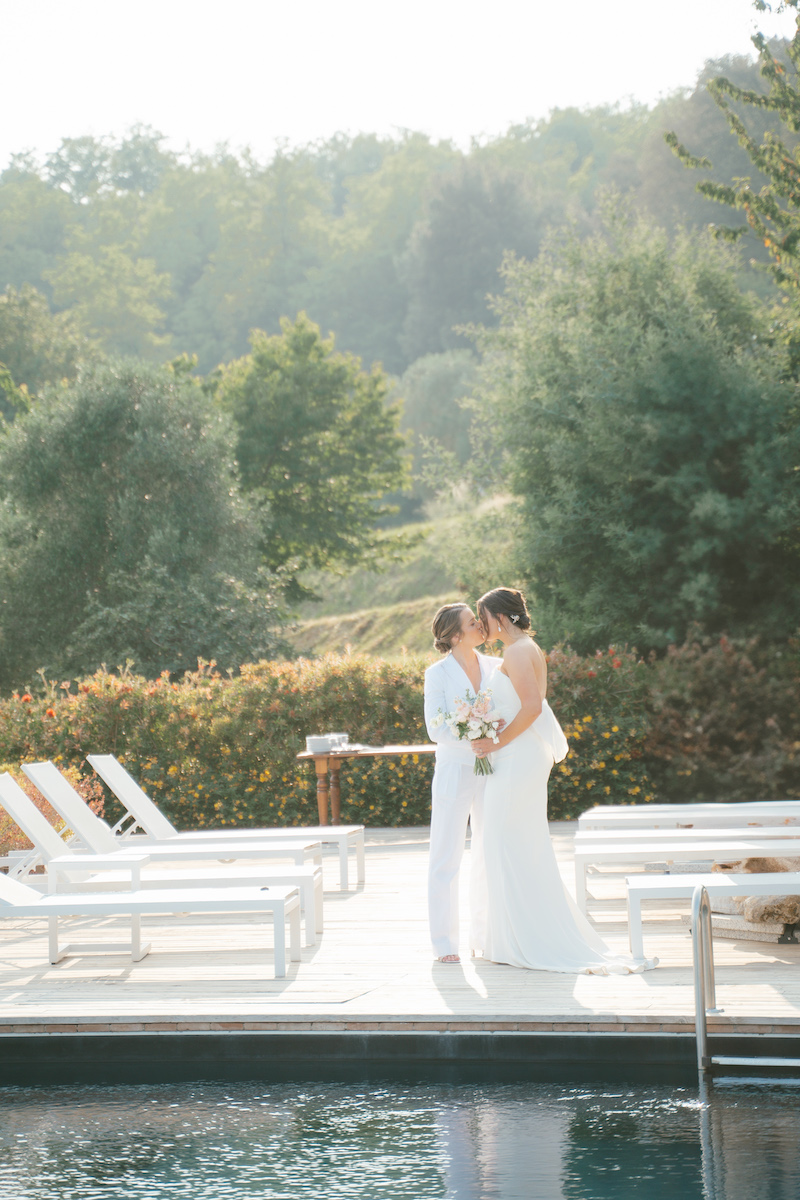 Two brides stand together by a poolside, one in a white suit and the other in a white dress, holding a bouquet with greenery in the background.