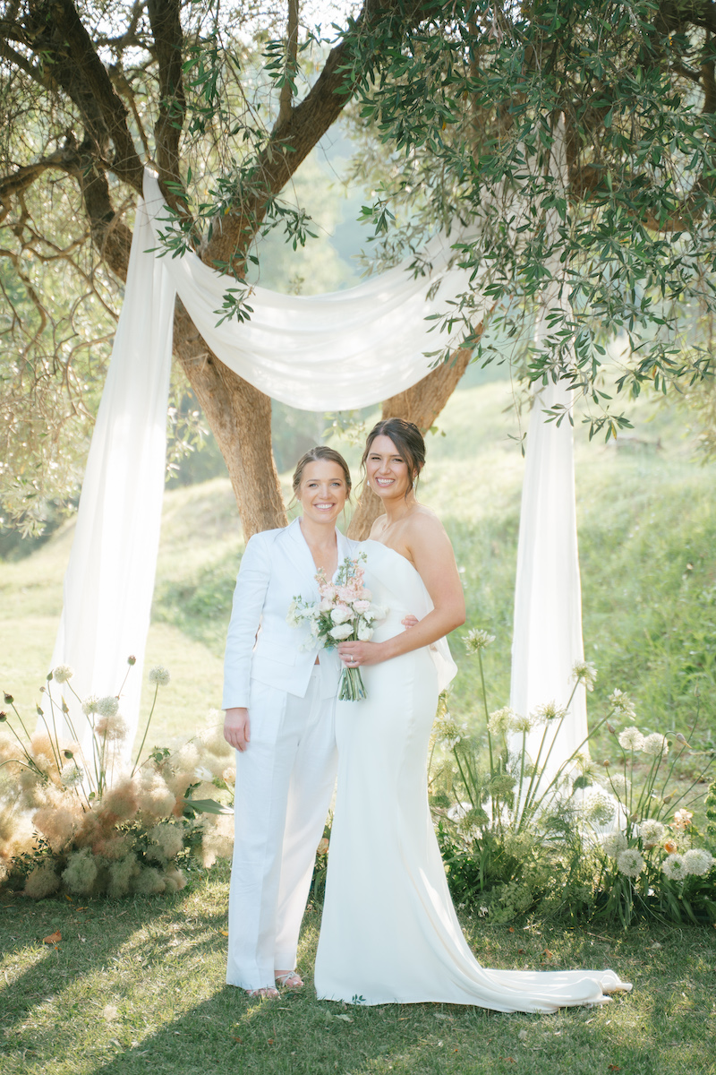 Two women in wedding attire stand and smile under a tree decorated with white fabric and flowers, holding a bouquet in an outdoor setting.