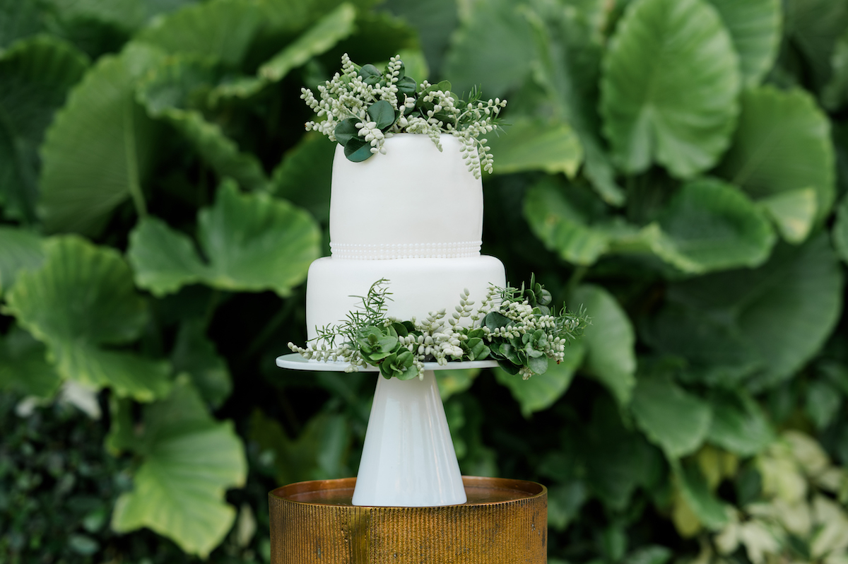 A two-tiered white cake decorated with green foliage and small white flowers, displayed on a white stand against a backdrop of large green leaves.
