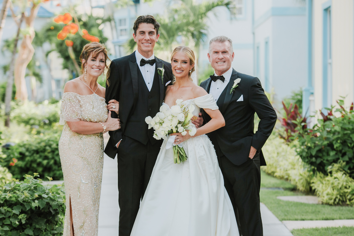 A bride and groom in formal wedding attire pose outdoors with two adults, all smiling at the camera, surrounded by greenery and buildings.