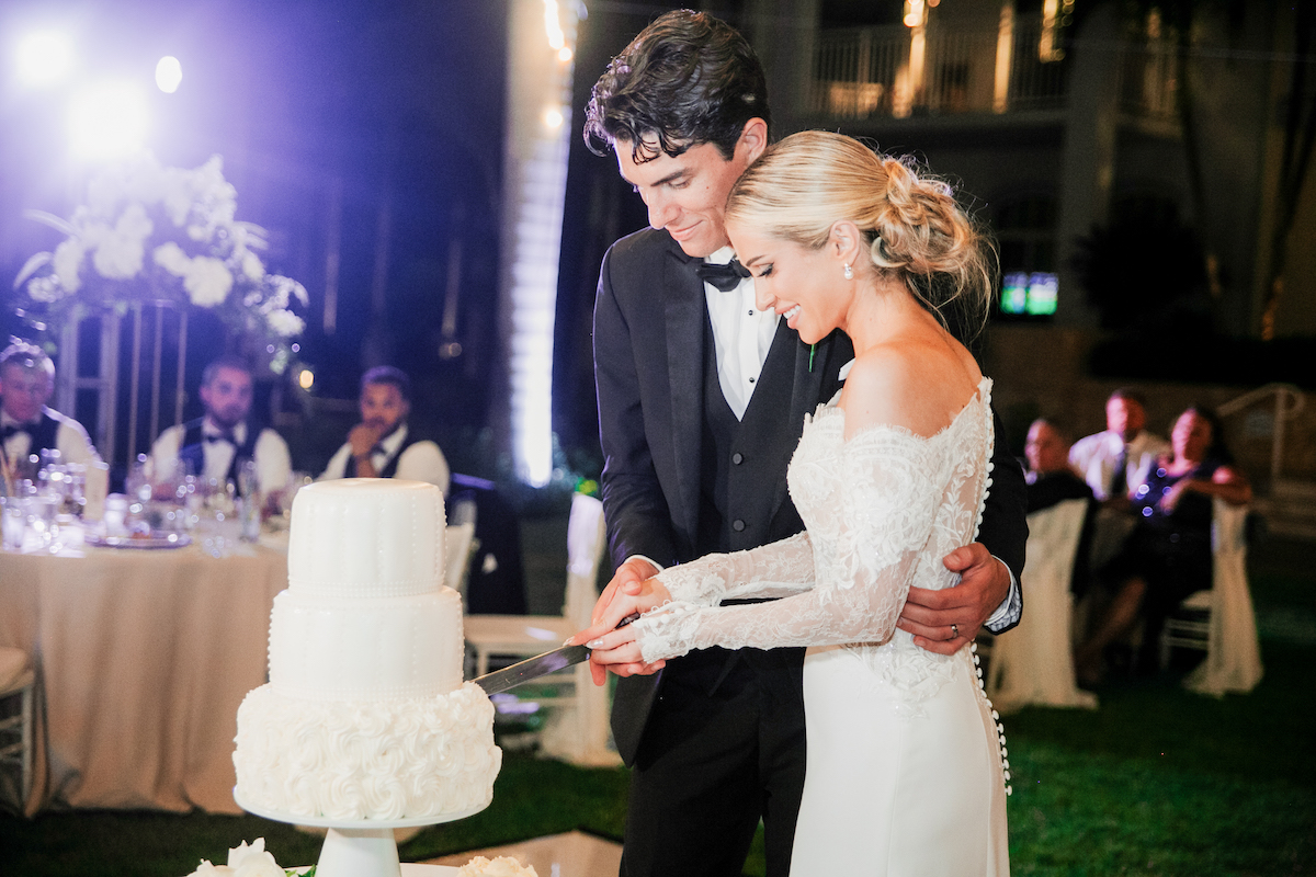 A bride and groom in formal attire cut their wedding cake together at a reception with guests seated in the background.
