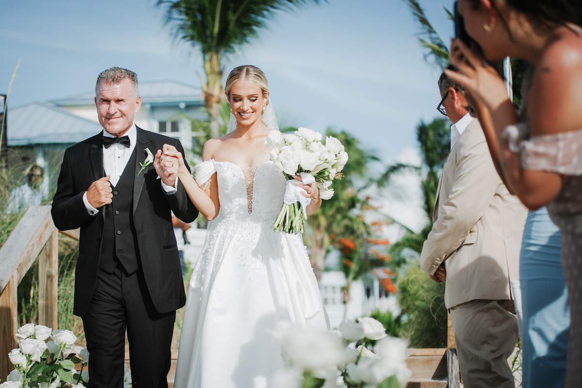A bride in a white gown walks arm-in-arm with an older man in a tuxedo outdoors, while a guest takes a photo and others stand nearby among flowers.