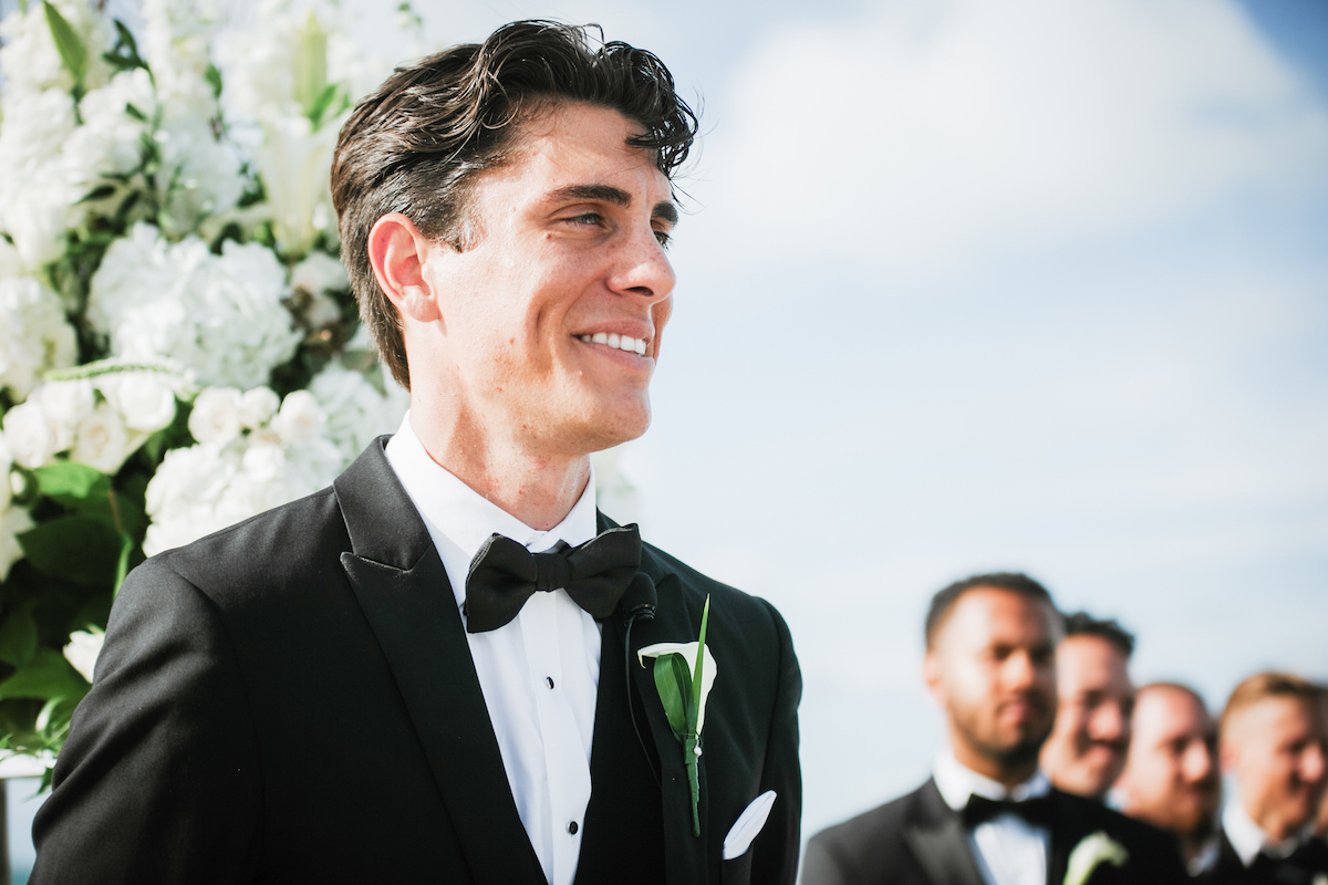 A man in a black tuxedo and bow tie stands smiling outdoors, with groomsmen and floral arrangements visible in the background.