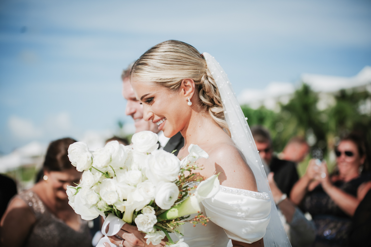 A bride in a white dress and veil holds a bouquet of white flowers, smiling outdoors with guests in the background.