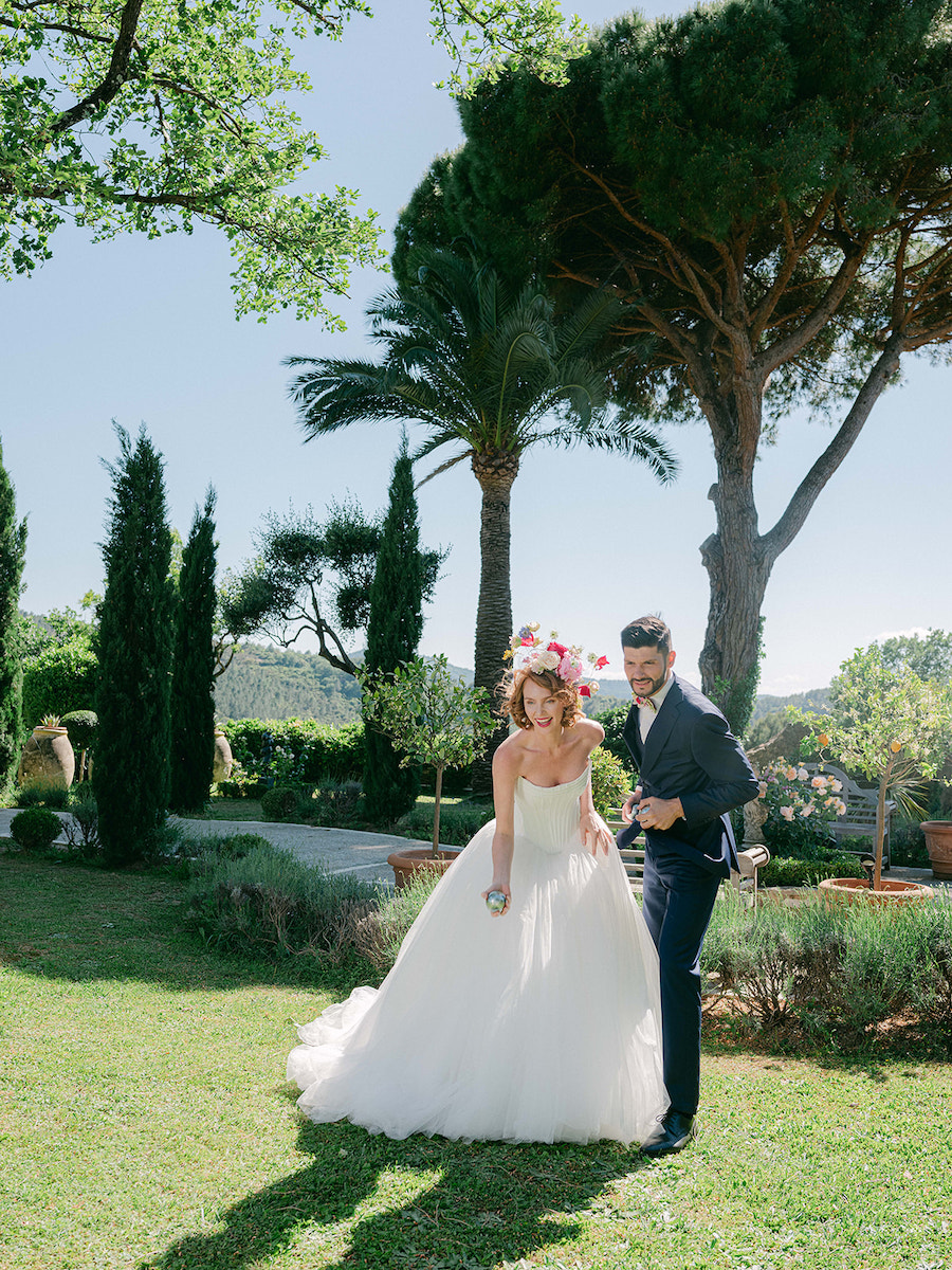 A bride in a white gown and a groom in a dark suit stand together on a grassy lawn with trees and greenery in the background.