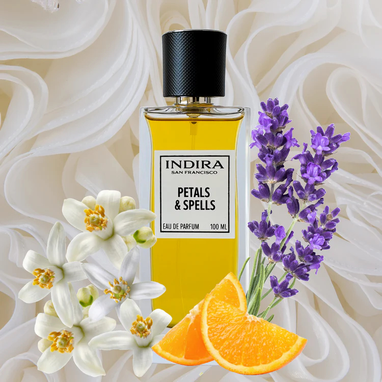 A bottle of Indira Petals &amp; Spells perfume is displayed with white flowers, lavender sprigs, and orange slices in front of a white, wavy textured background.