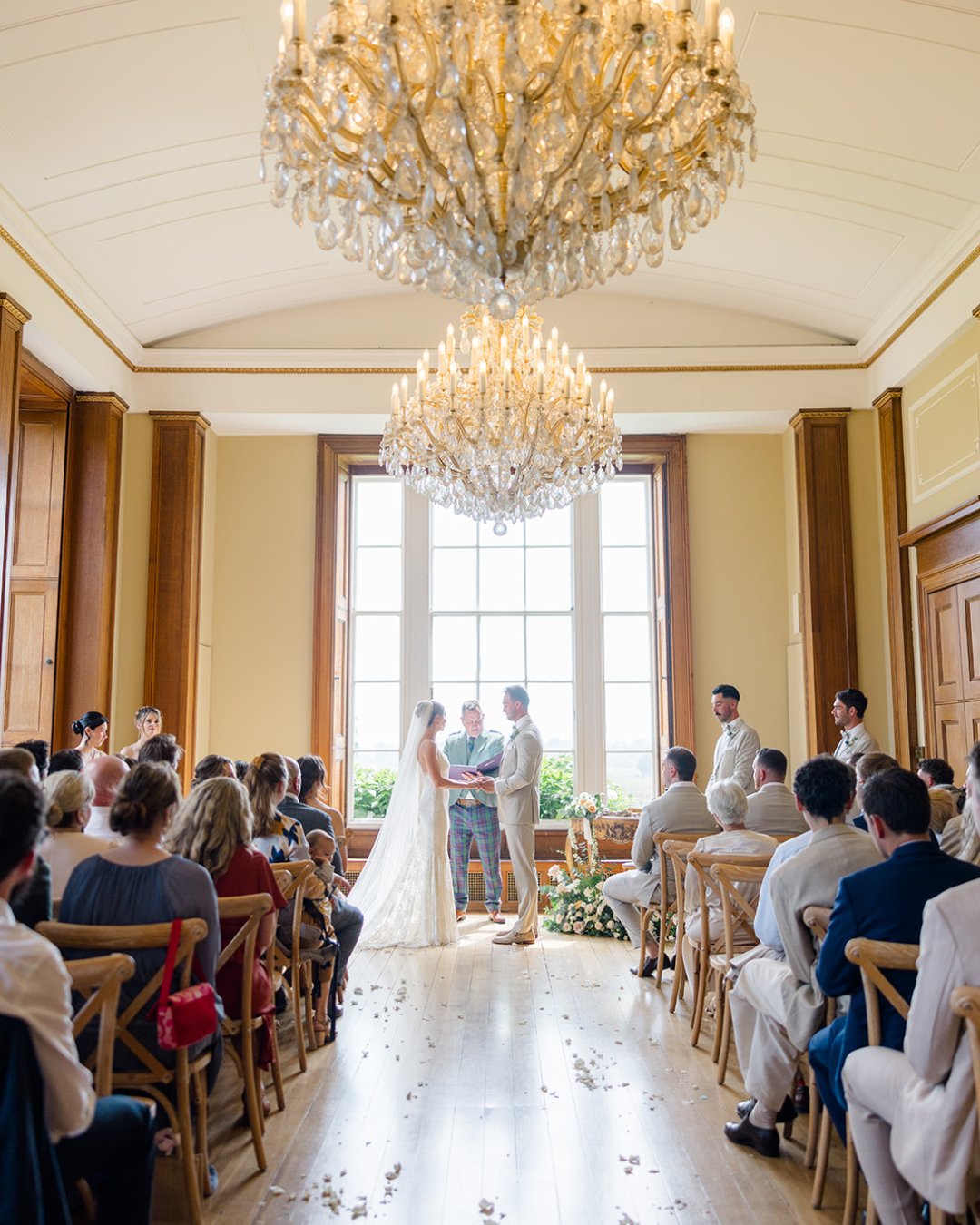 A bride and groom stand at the altar in a sunlit room with chandeliers, surrounded by guests seated on wooden chairs during a wedding ceremony.