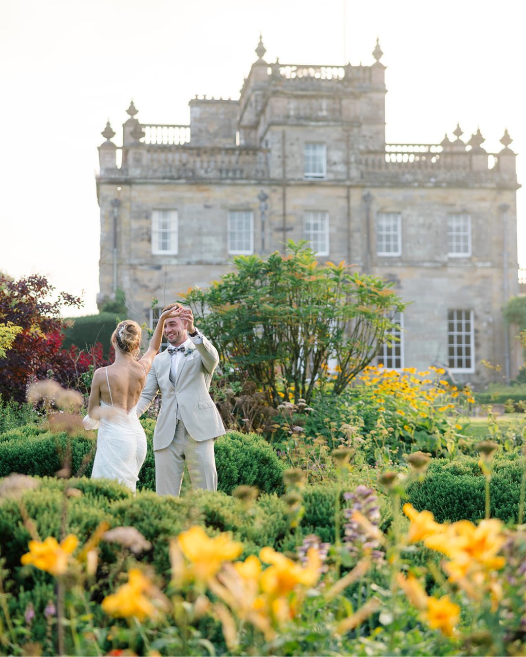 A bride and groom stand in a garden, with a historic stone building in the background. The groom appears to be waving or gesturing, while the bride looks on.
