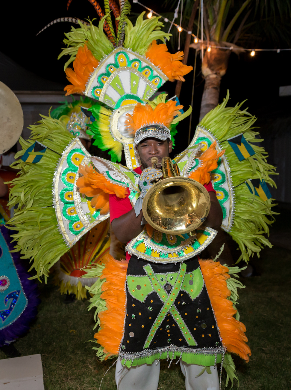 A Junkanoo performer in a bright, elaborate feathered costume plays a brass instrument outdoors at night.