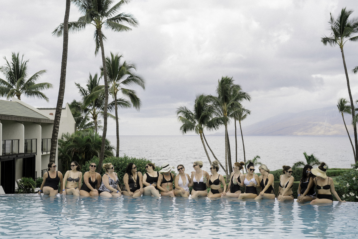 A group of women in swimsuits sit on the edge of a pool with their feet in the water, with palm trees and the ocean in the background.