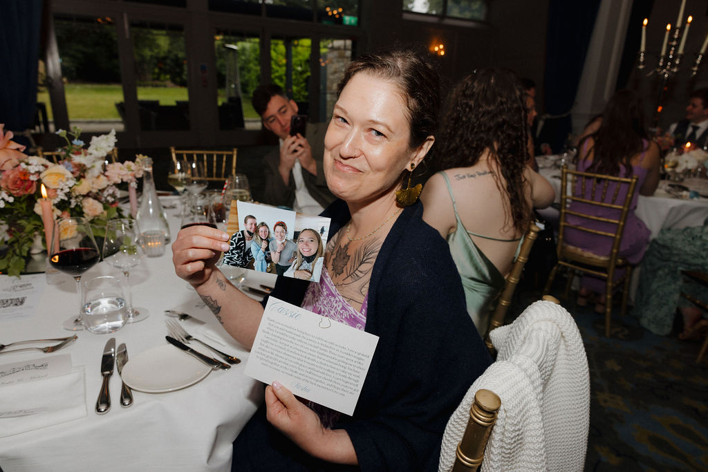 A woman seated at a dinner table holds up a photograph and a handwritten letter, smiling at the camera. Other guests and floral arrangements are visible in the background.