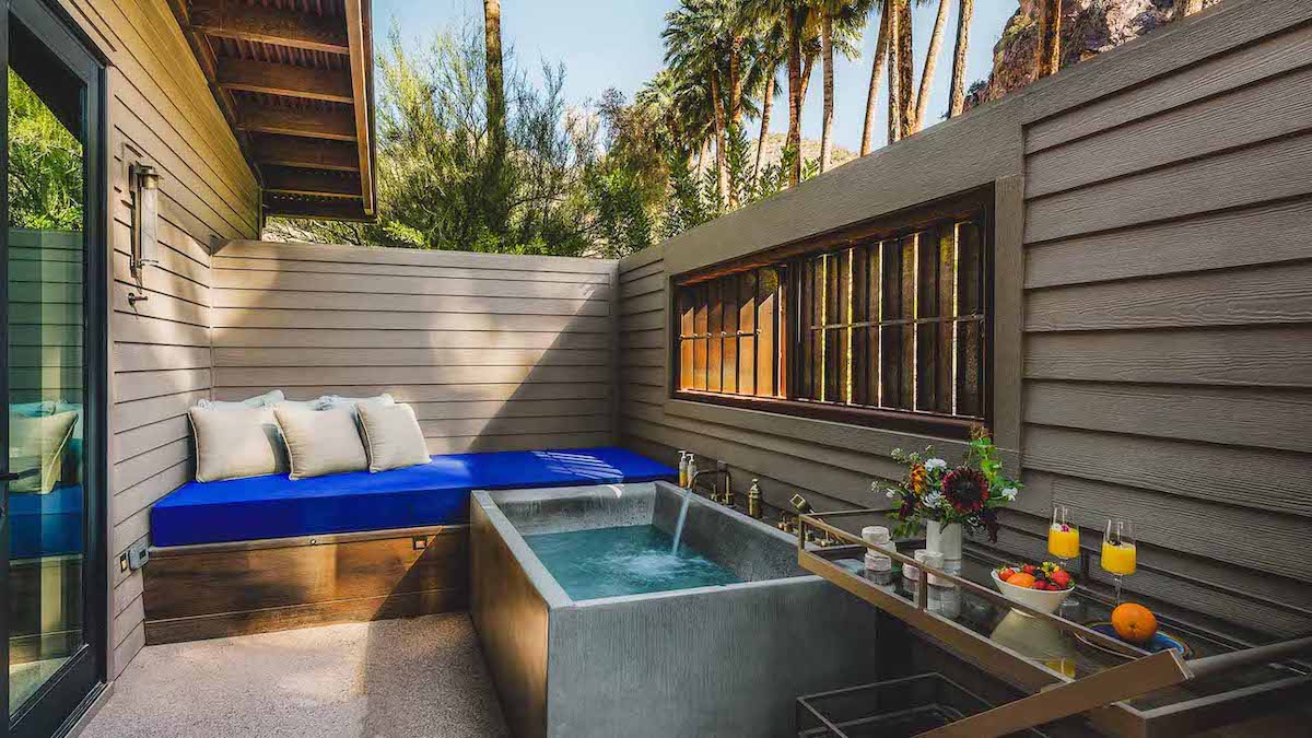 Outdoor patio with a small concrete soaking tub, cushioned bench with blue pillows, and a table holding drinks and flowers, surrounded by wooden walls and greenery.