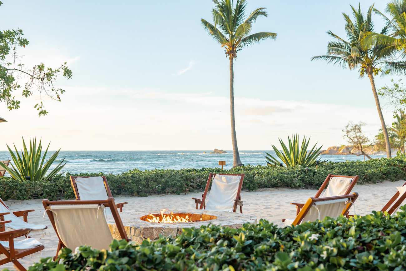 Wooden lounge chairs surround a fire pit on a sandy beach with palm trees, overlooking the ocean under a clear sky.