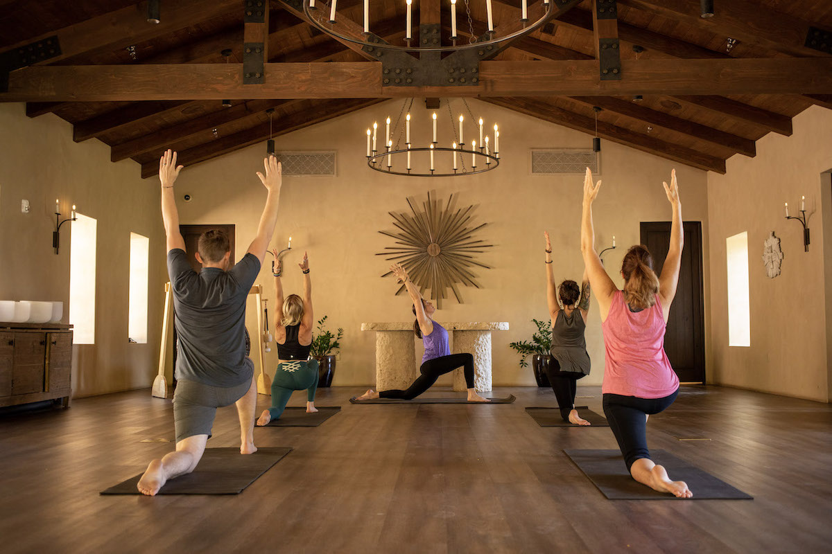 Five people practice yoga on mats in a spacious room with wooden floors, high ceilings, chandeliers, and warm light. They are in a kneeling lunge pose with arms raised.