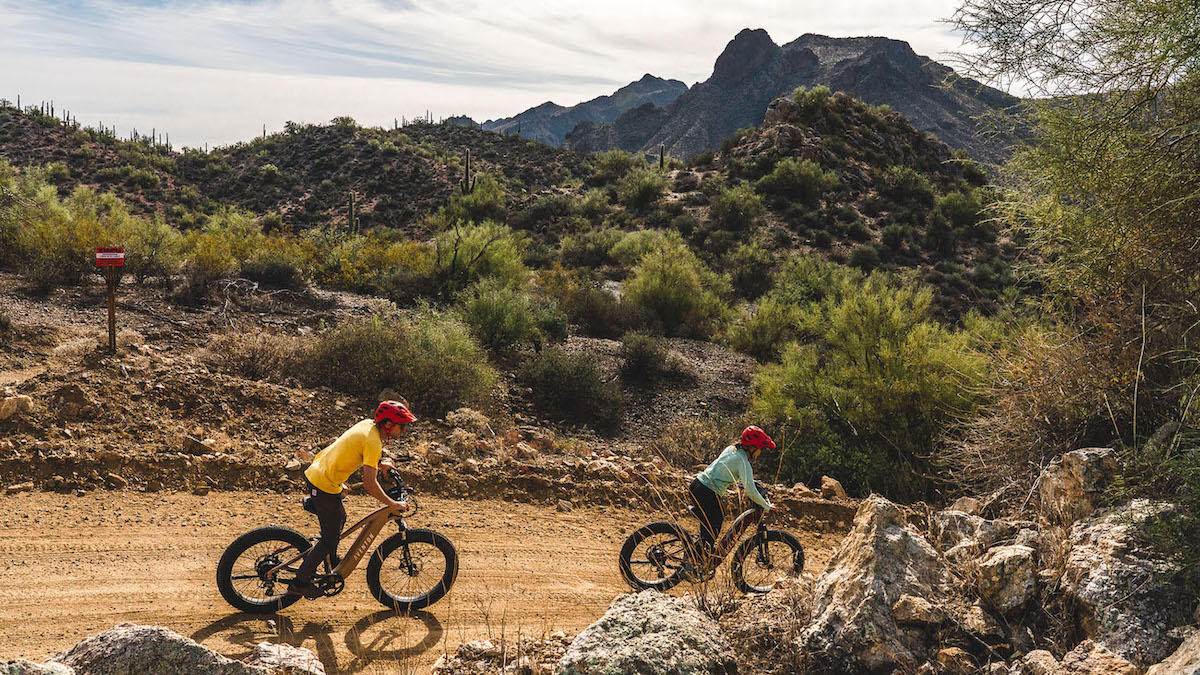 Two people wearing helmets ride bicycles on a dirt trail through a desert landscape with mountains and shrubs in the background.