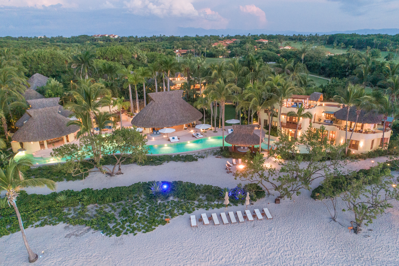 Aerial view of a beachfront resort with thatched-roof buildings, a pool, lounge chairs, palm trees, and white sand, surrounded by lush greenery.
