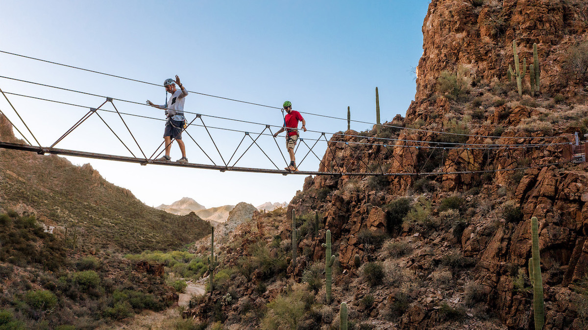 Two people wearing helmets cross a narrow suspension bridge over a rocky canyon with cacti and desert landscape in the background.