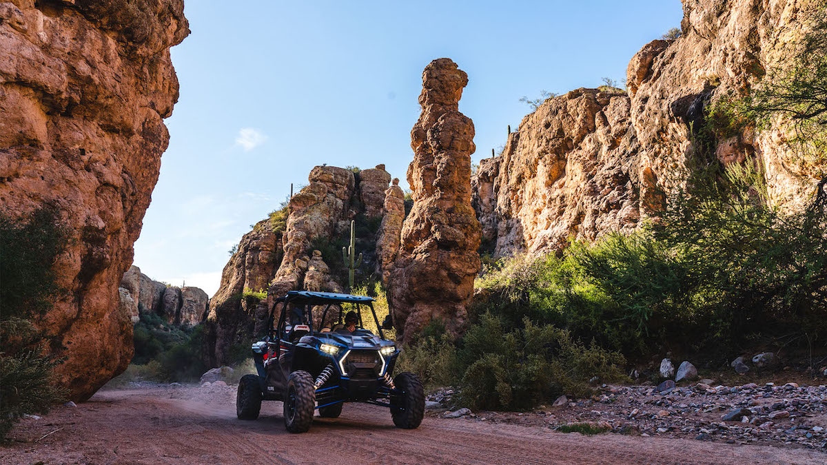 An off-road vehicle drives on a dirt path between tall, rocky canyon walls under a clear blue sky.
