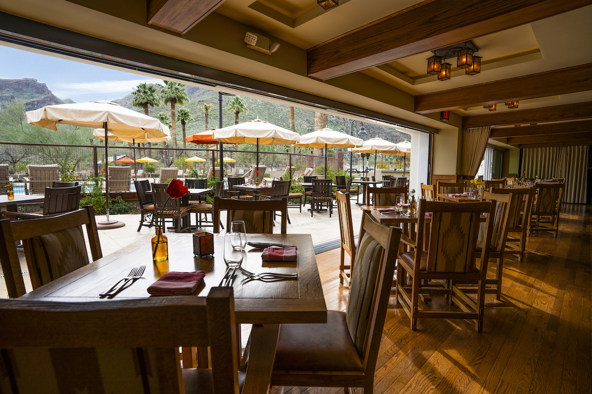 Indoor restaurant seating with wooden tables and chairs, set for dining, overlooks an outdoor patio area with umbrellas and mountains in the background.