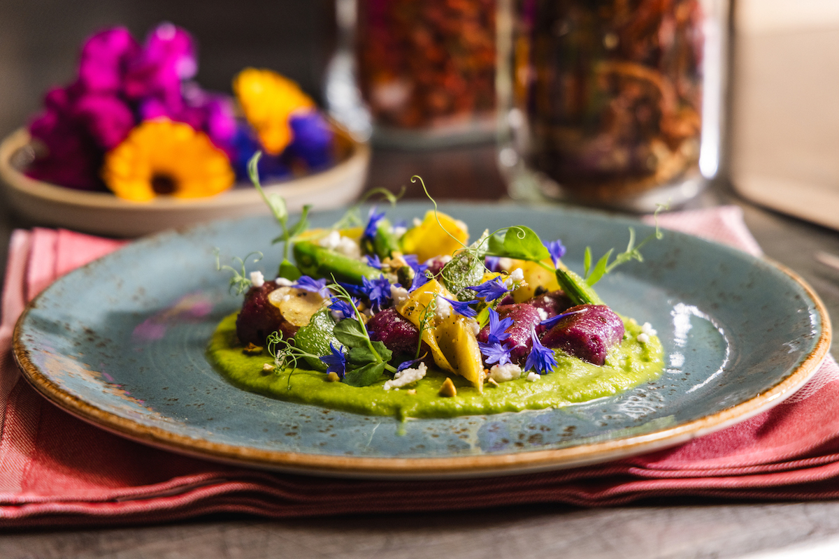 A blue ceramic plate with a colorful dish featuring assorted vegetables, edible flowers, and a green sauce, set on a pink napkin with blurred flowers and jars in the background.