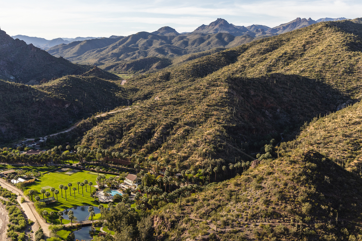 Aerial view of a desert landscape with rugged mountains and a green area with palm trees and ponds in the foreground.