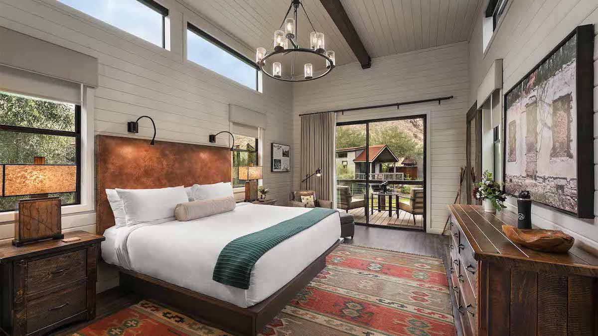 A spacious bedroom with a king-sized bed, rustic wooden furniture, large windows, a chandelier, colorful rug, and a sliding glass door leading to an outdoor patio.