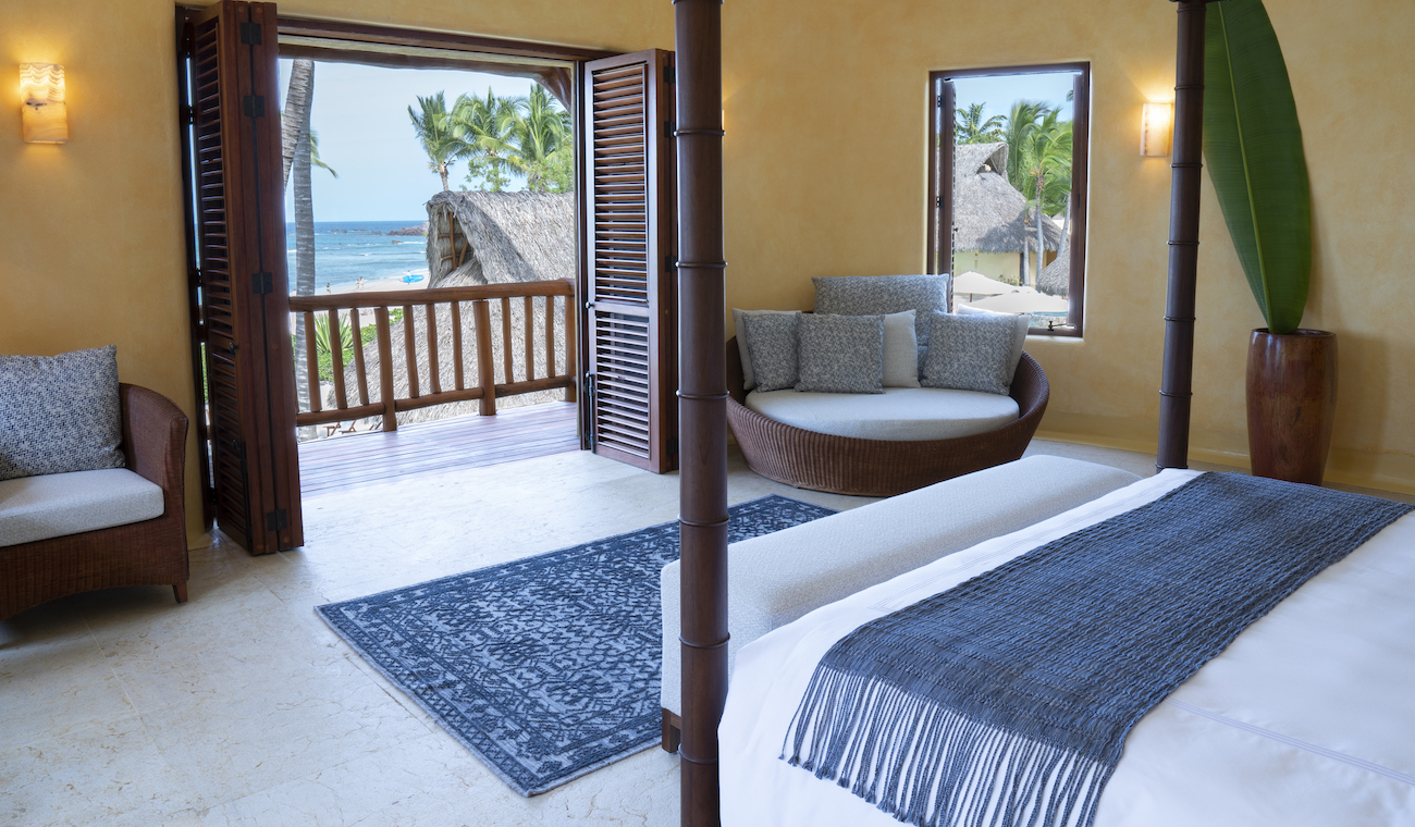 A resort room with a bed, seating area, large windows, open wooden doors leading to a balcony, and a view of the ocean and palm trees outside.