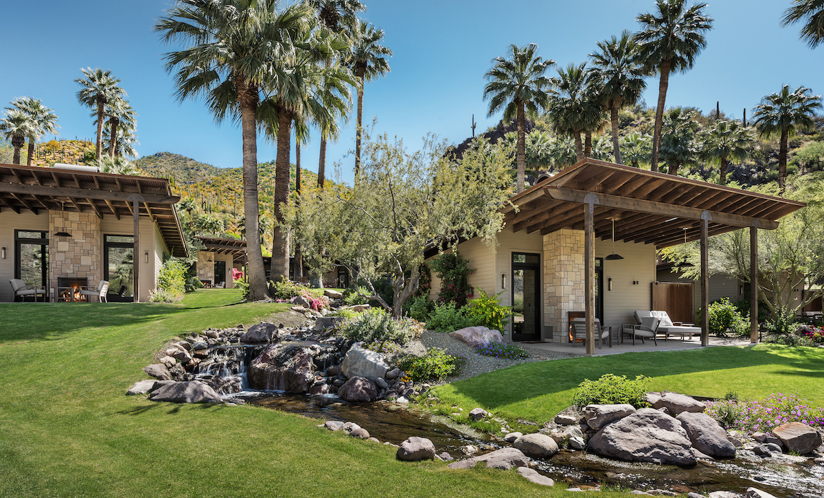 A landscaped yard with a small stream, rocks, two modern stone buildings, palm trees, and mountains in the background under a clear blue sky.