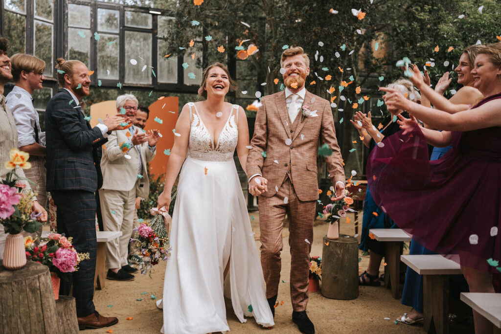 A bride and groom walk down the aisle smiling as guests throw confetti in celebration at an indoor wedding ceremony.