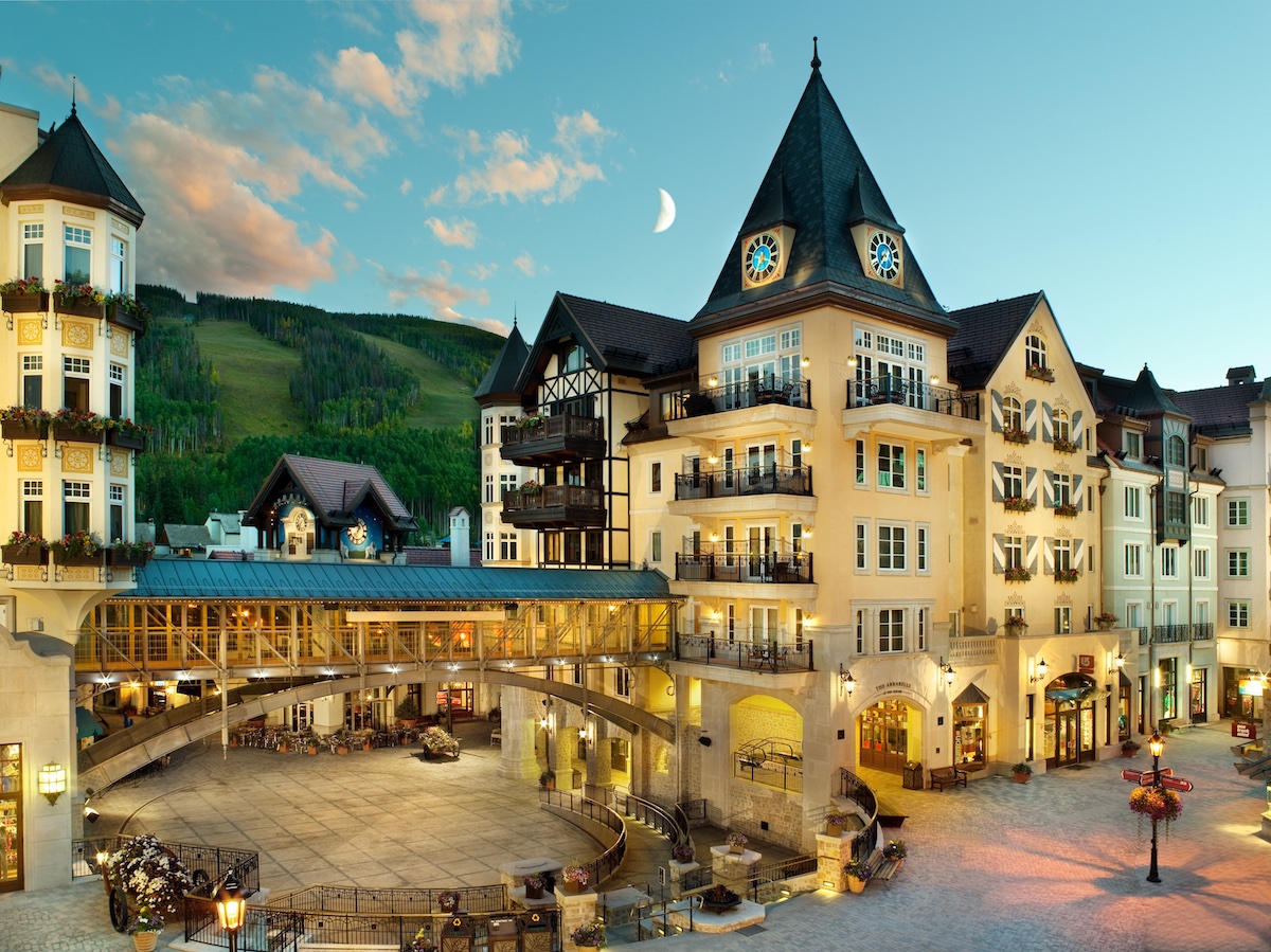 A European-style village square with ornate buildings, a clock tower, balconies, and a mountain backdrop under a clear sky at dusk.
