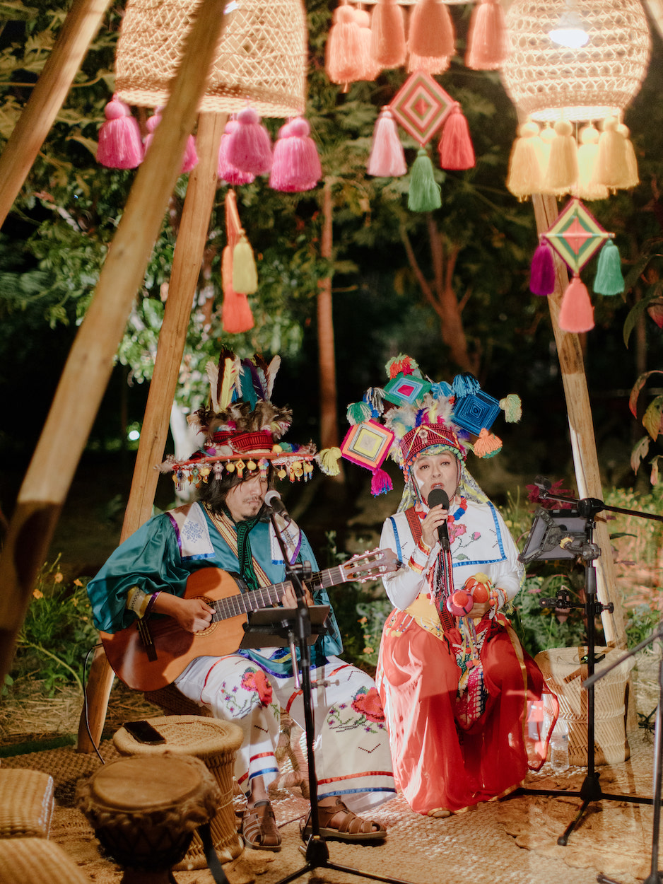 Two performers in colorful traditional attire play guitar and sing under a canopy decorated with woven lamps and tassels at an outdoor event.