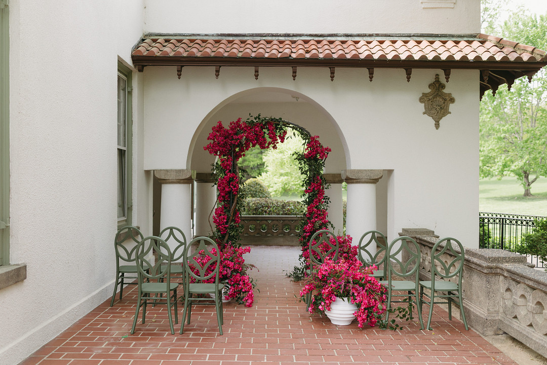 A small outdoor wedding ceremony setup with green chairs and an archway decorated with pink flowers on a brick patio.