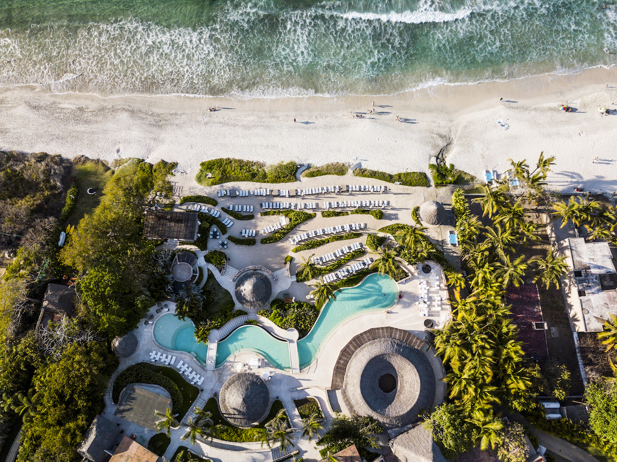 Aerial view of a beachfront resort with multiple pools, lounge chairs, palm trees, and direct access to a sandy beach and ocean waves.