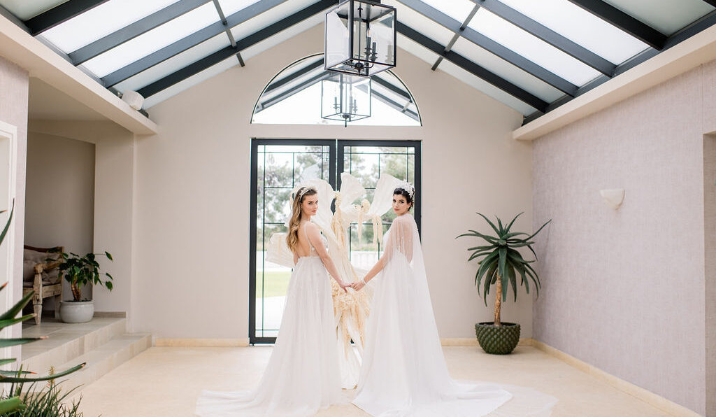 Two women in white gowns stand indoors, holding hands and facing the camera, with a large window, sculpture, and potted plant behind them.