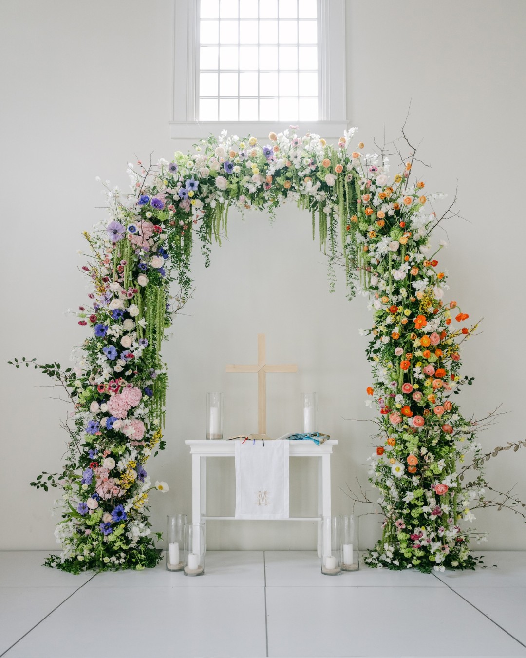 A floral arch with multicolored flowers frames a small white table with candles and a cross, set against a white wall with a window above.