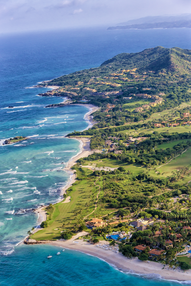 Aerial view of a lush green coastline with beaches, waves, and scattered buildings along the shore under a clear sky.