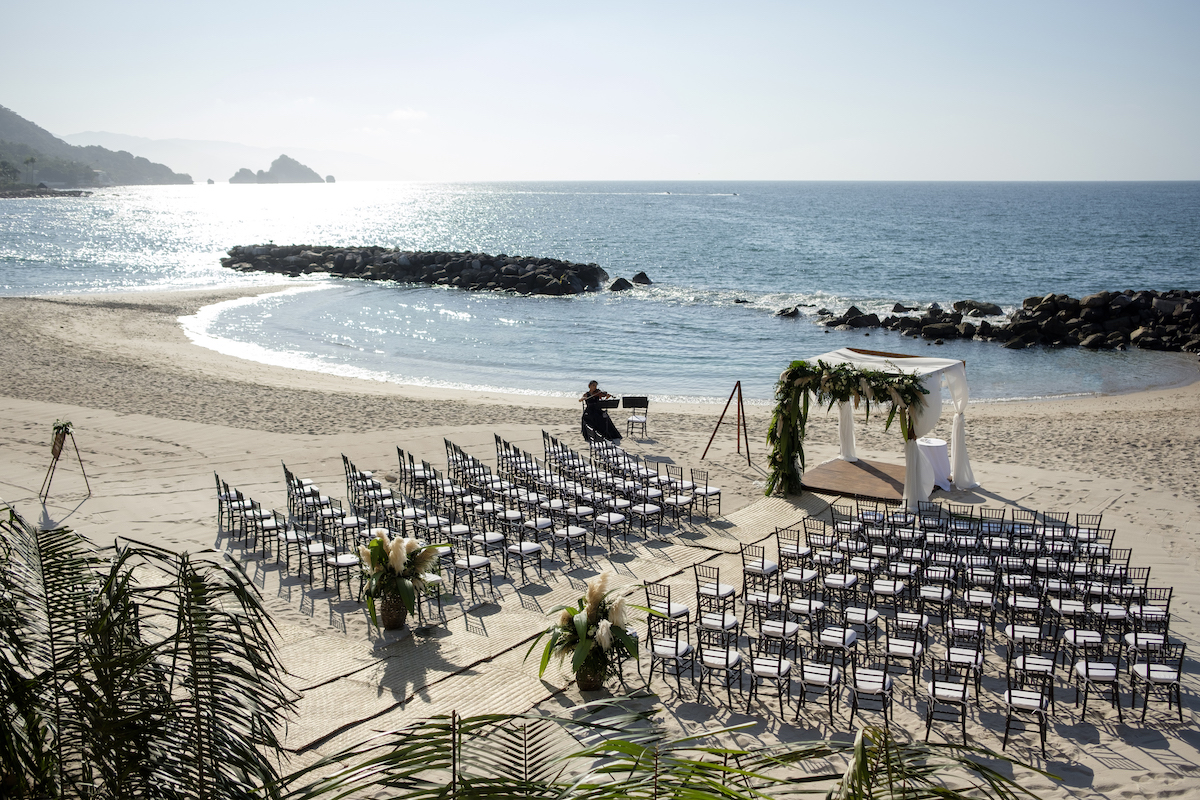 Rows of chairs are arranged on a sandy beach facing an archway decorated for a wedding ceremony, with the ocean and rocks in the background.