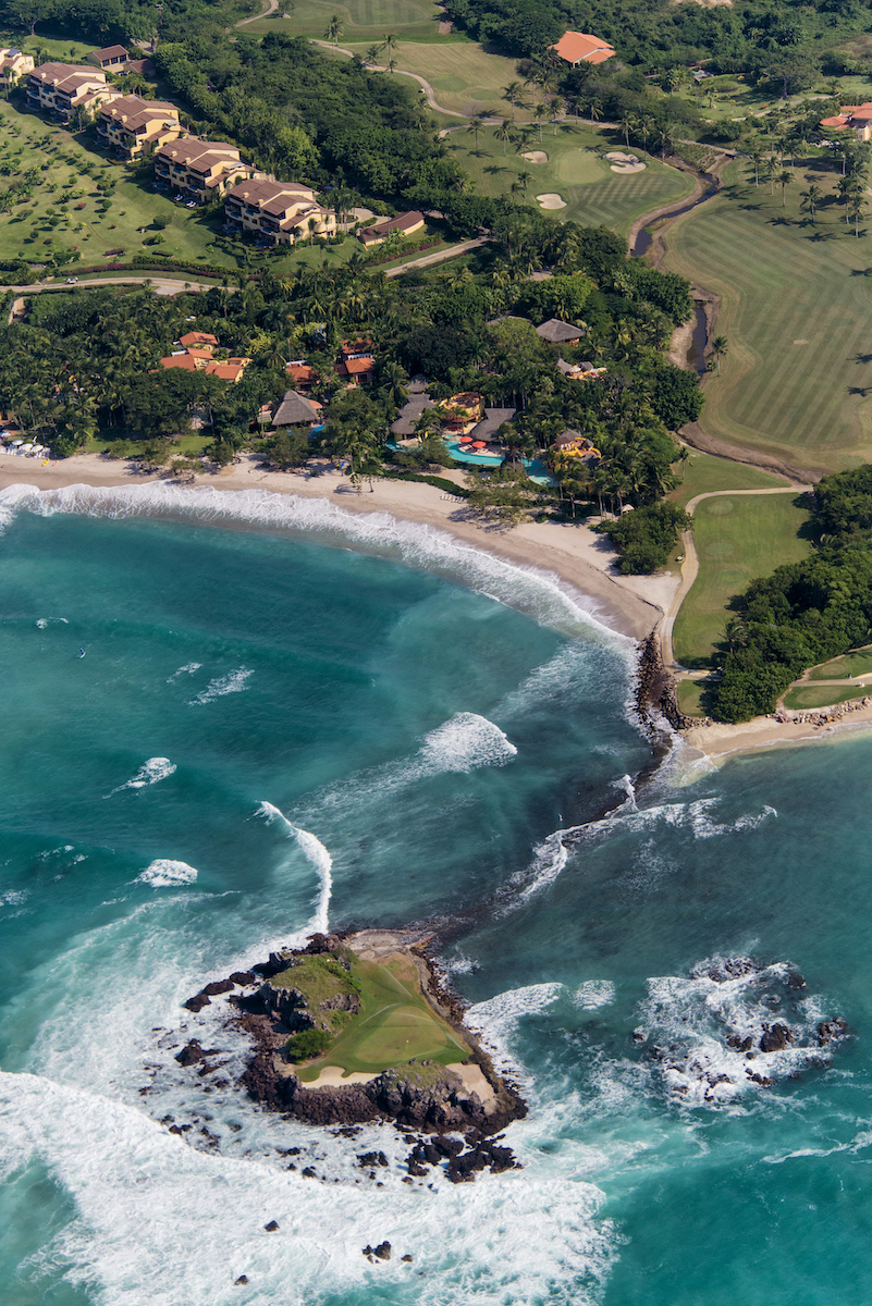 Aerial view of a coastal area with turquoise waves, sandy beach, rocky outcrop, and nearby houses surrounded by greenery.