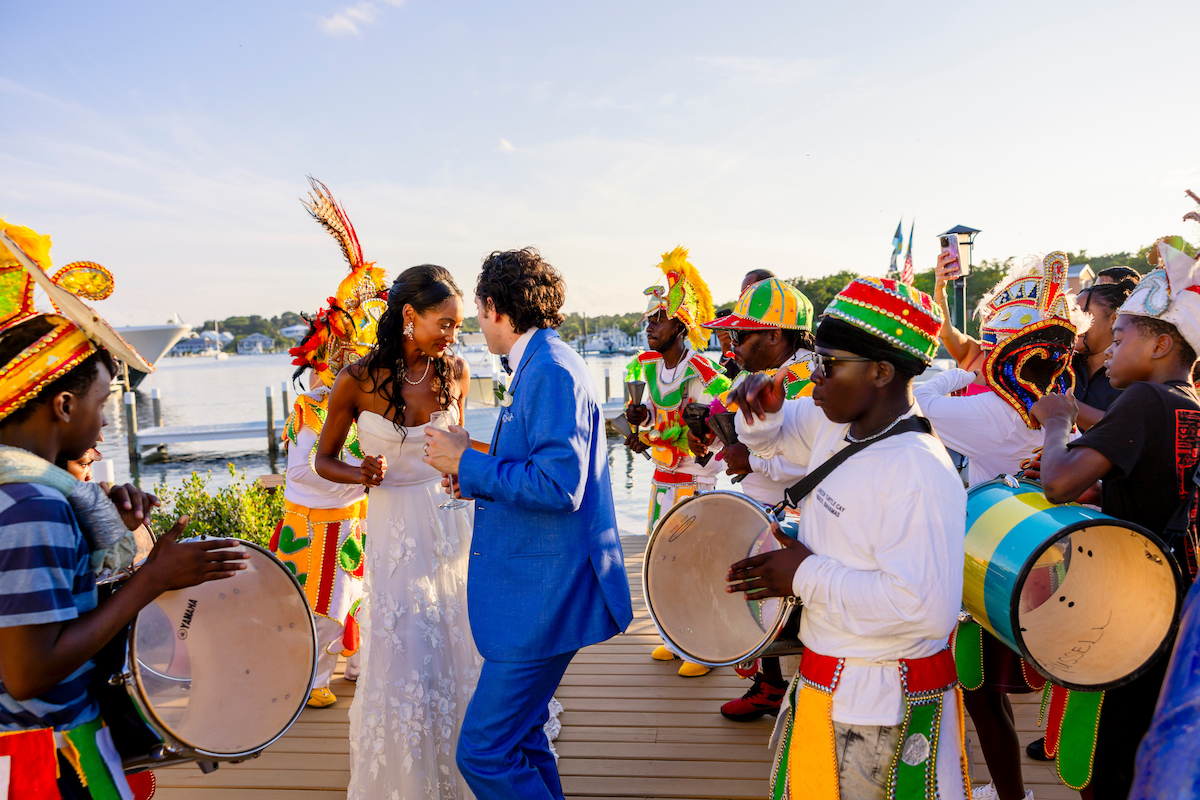 A bride and groom dance outdoors, surrounded by musicians in colorful costumes playing drums near a waterfront.